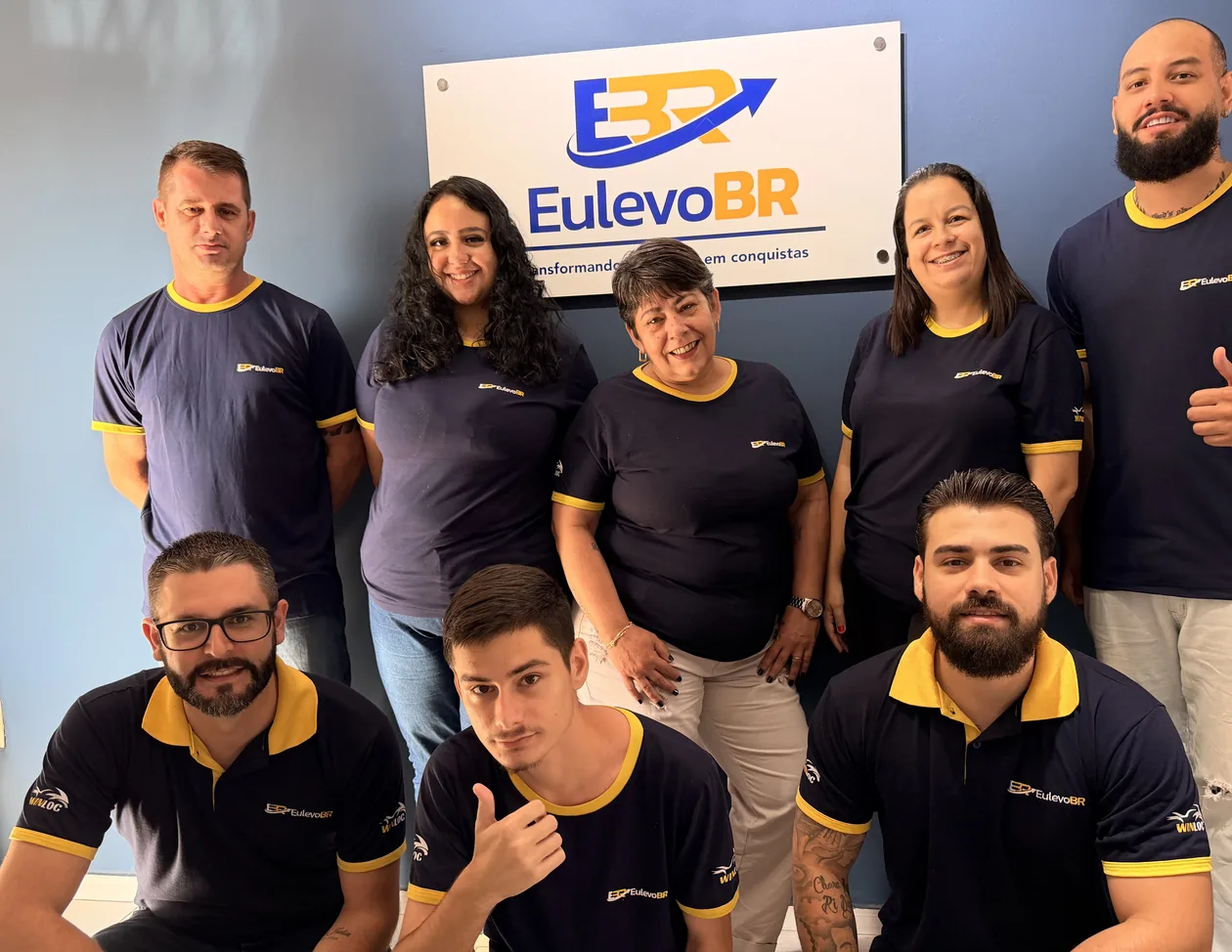 Equipe da EuLevoBR reunida na sede, de uniforme, embaixo da placa da marca